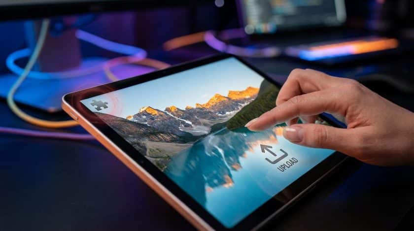 Hand interacts with sleek tablet displaying mountain landscape, upload button, and a puzzle piece icon with a red glow.