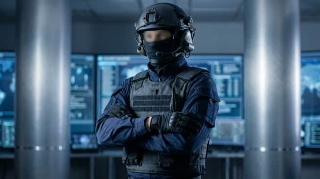 Anonymous figure in dark tactical gear, helmet, and headset, arms crossed, in a modern, blurred command center with digital screens.