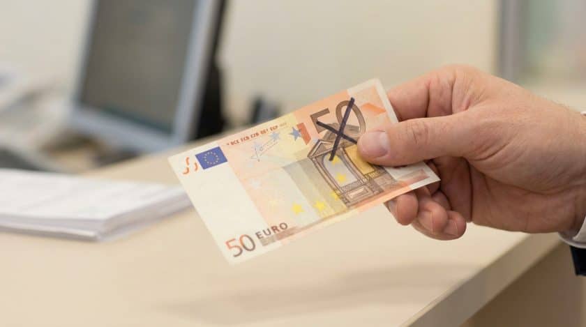 A clean hand gently holds a 50 euro banknote marked with a black felt-tip 'X', set against a blurred professional desk background.