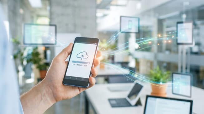 Main tenant un smartphone affichant un écran d'upload cloud à 90%. Des flux de données lumineux connectent l'appareil à d'autres écrans en arrière-plan.